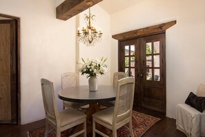 A dining area with a round table, four chairs, a chandelier, a vase with flowers, and a wooden door.
