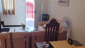 Un salon avec un canapé, une table à manger et une chaise. Il y a une fenêtre avec des rideaux, une horloge au mur et une porte qui donne sur l’extérieur.