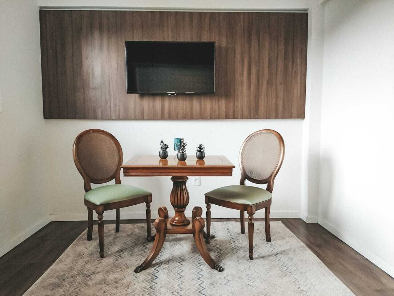 A dining area with a wooden table and two chairs, a flat-screen TV mounted on the wall, and a patterned rug on the floor.