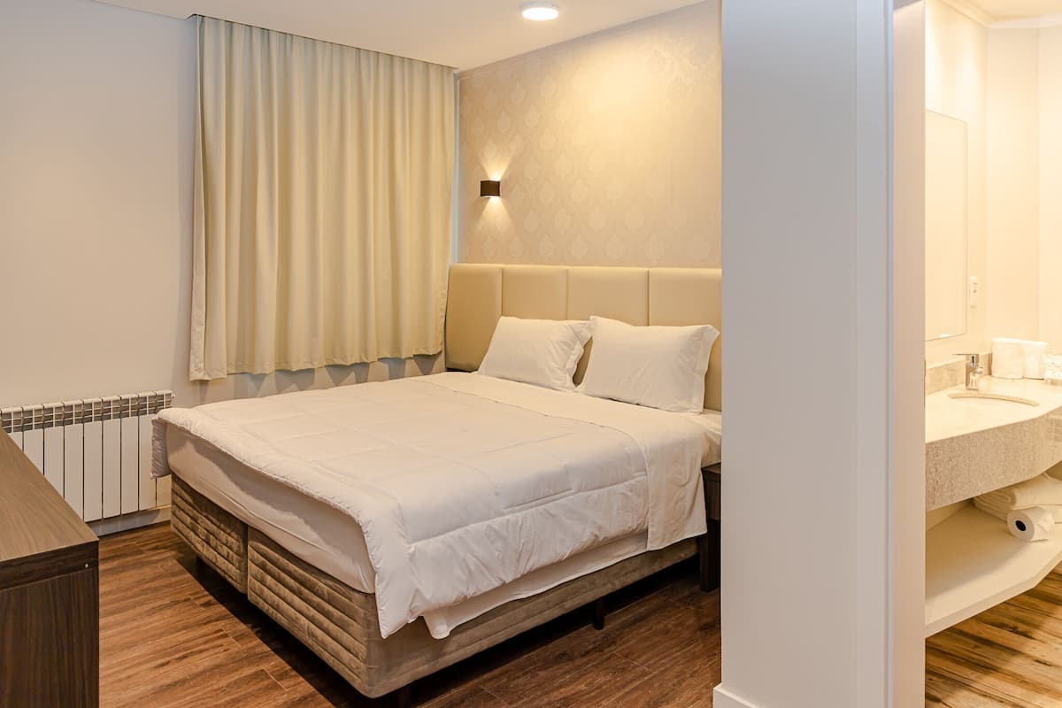 Habitación de hotel con cama, mesita de noche, luz en la pared, radiador y baño con lavabo y espejo.