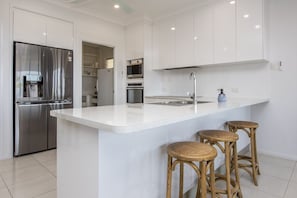 Eine moderne Küche mit weißen Schränken, einem Edelstahlkühlschrank, einem eingebauten Backofen und einer zentralen Kochinsel mit Sitzgelegenheiten.