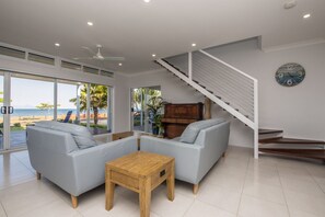 Ein modernes Wohnzimmer mit einer Sofagarnitur, einem Holztisch, einer Treppe und großen Fenstern mit Blick auf den Strand.
