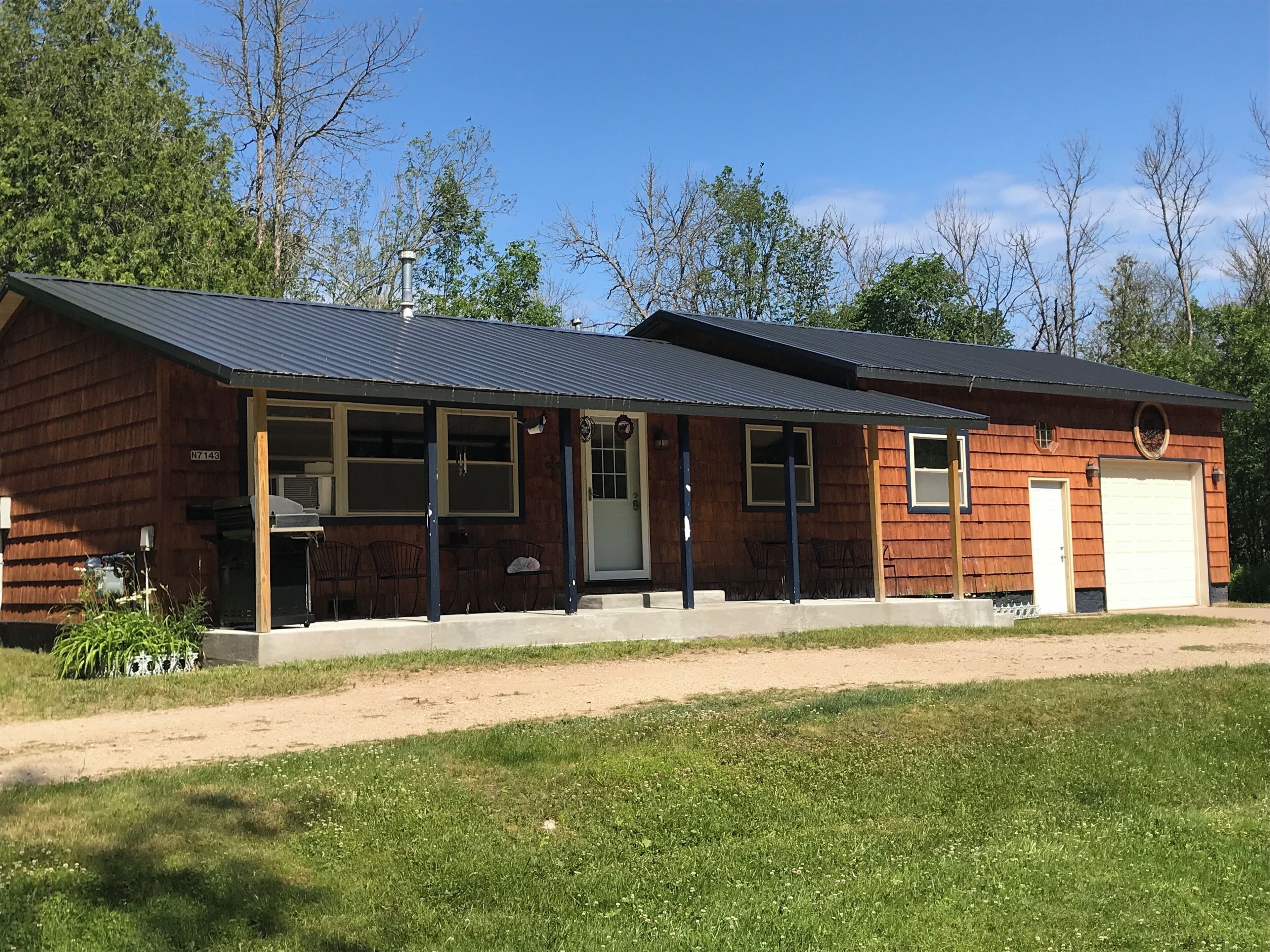 Une cabane en bois à deux étages, avec un toit en métal, une véranda à l’avant dotée de chaises et un garage.