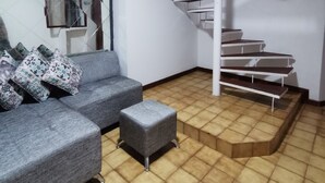 Ein modernes Wohnzimmer mit einem grauen Sofa, gemusterten Kissen, einem kleinen Hocker und einer Wendeltreppe mit Holzstufen.