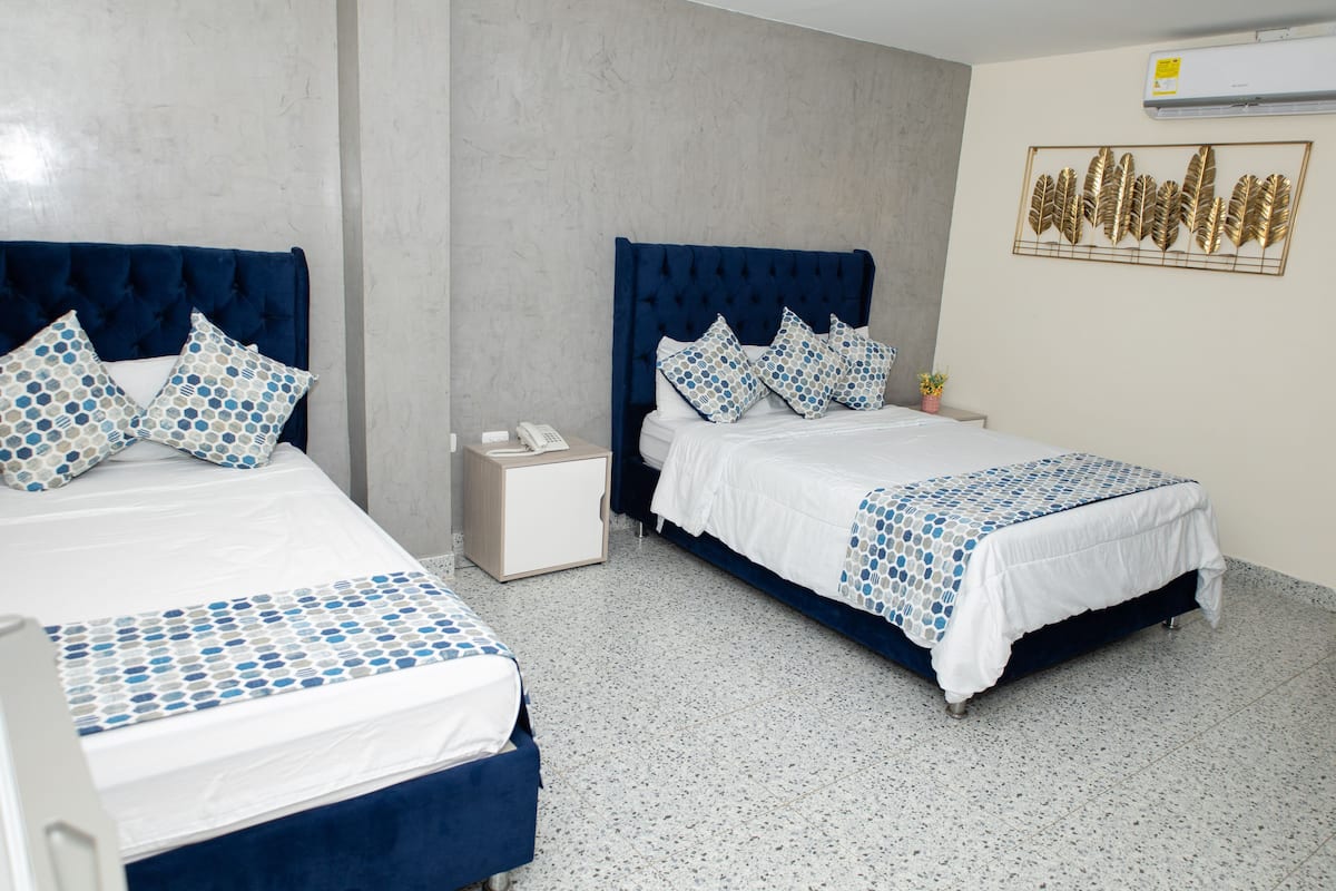 Une chambre d’hôtel avec deux lits, chacun doté d’une tête de lit bleue et de draps blancs. On y trouve une petite table de chevet avec un téléphone et une lampe. La chambre a un sol carrelé et un climatiseur fixé au mur.