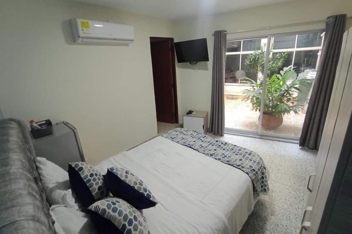 Une chambre d’hôtel avec un lit, une télévision, un climatiseur, une fenêtre avec des rideaux et une plante en pot à l’extérieur.
