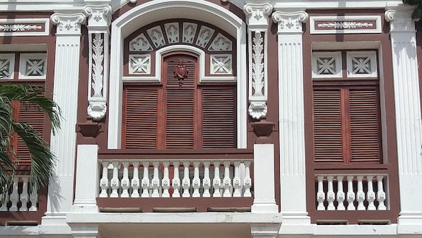 Ein Gebäude mit einer symmetrischen Fassade, das ein zentrales Rundbogenfenster mit Fensterläden und zwei Doppeltüren aufweist. Verzierte Gesimse und ein Balkon mit Brüstung sind vorhanden.