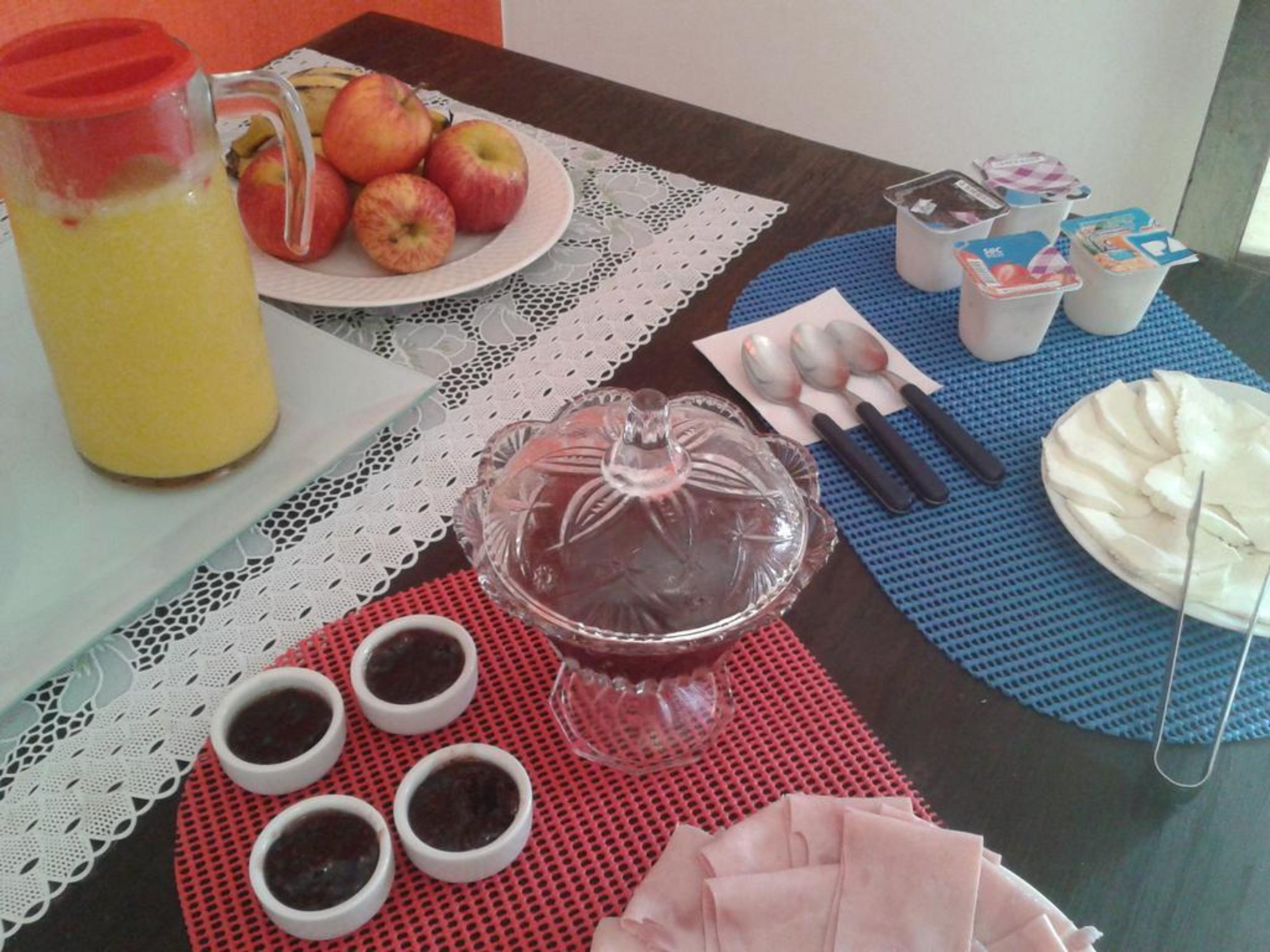 A table with a fruit plate, a pitcher of juice, a glass cake stand, small bowls of dip, and a plate of sliced meat.