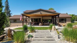 A two-story house with a covered patio, stone steps leading to the entrance, and a flag on the left. - Large 7BR Home Near Lake CDA | Backyard Oasis & Game Room (Coeur d'Alene)