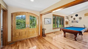 A room with a pool table, wooden flooring, a large window with a view of greenery, and a door with glass panels. - Large 7BR Home Near Lake CDA | Backyard Oasis & Game Room (Coeur d'Alene)