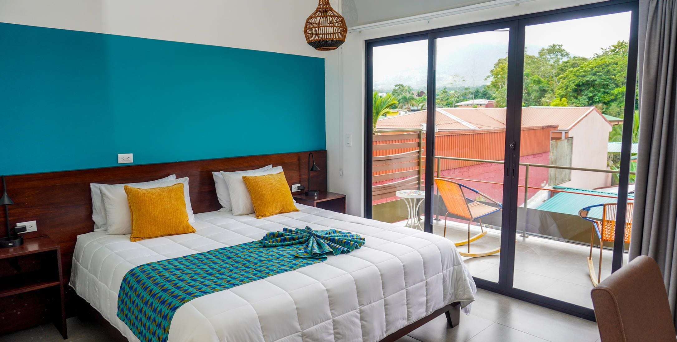 A hotel room with a large bed, a balcony with a table and chairs, and a view of buildings and greenery.