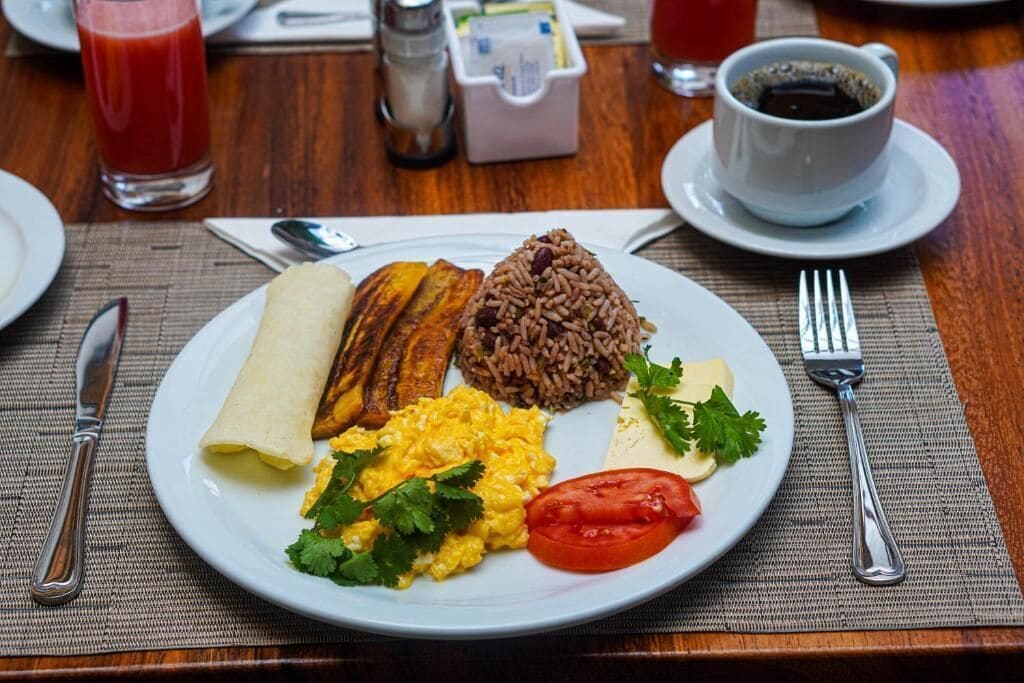 A plate of breakfast food including eggs, rice, plantains, and a wrap, with a cup of coffee and a glass of juice on the table.