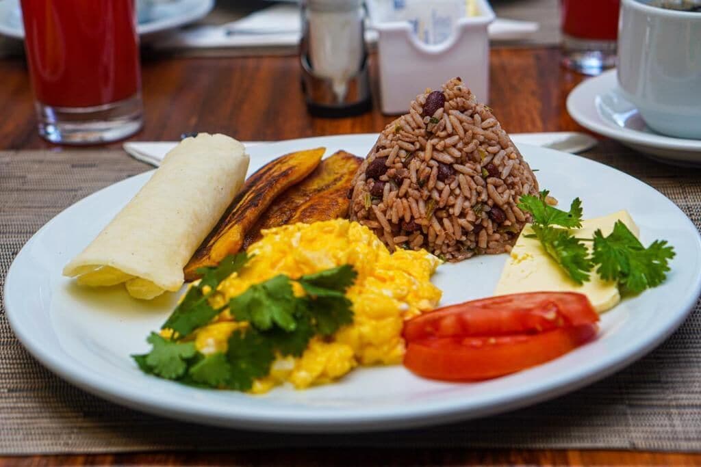 Un plato con una variedad de alimentos para el desayuno, incluyendo huevos revueltos, arroz, un omelette enrollado, plátanos fritos y tomate.