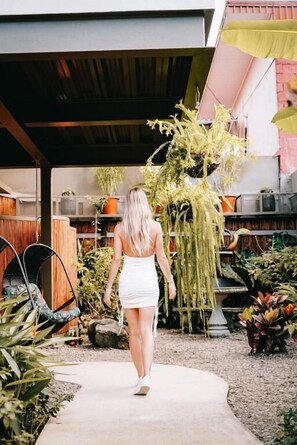 A woman walking on a concrete path in a garden area with various plants and a wooden fence. - La Fortuna Lodge by Treebu Hotels (La Fortuna)