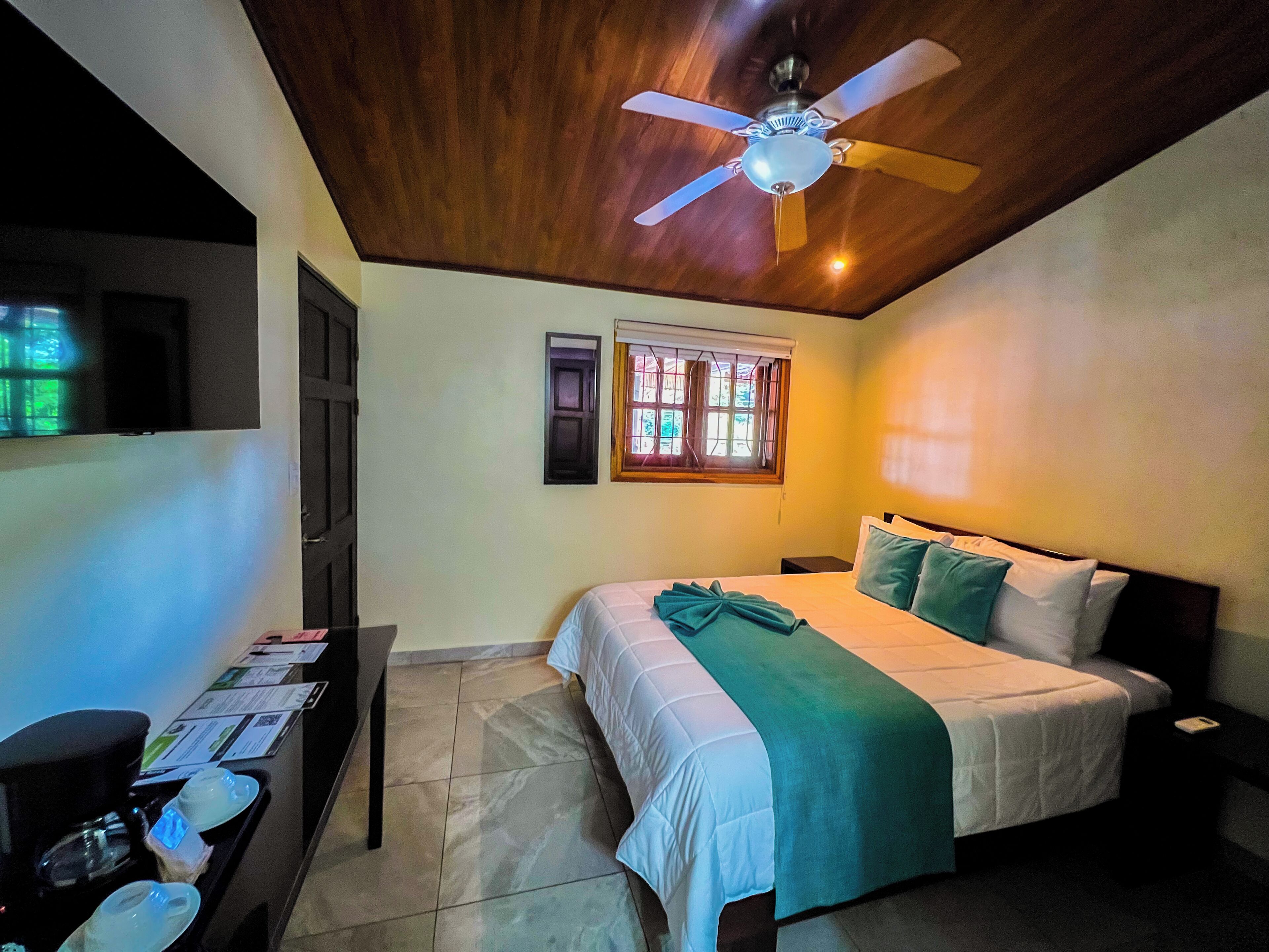 Une chambre à coucher avec un lit, un ventilateur de plafond, une fenêtre avec des volets et un bureau sur lequel se trouve un plateau contenant divers objets.