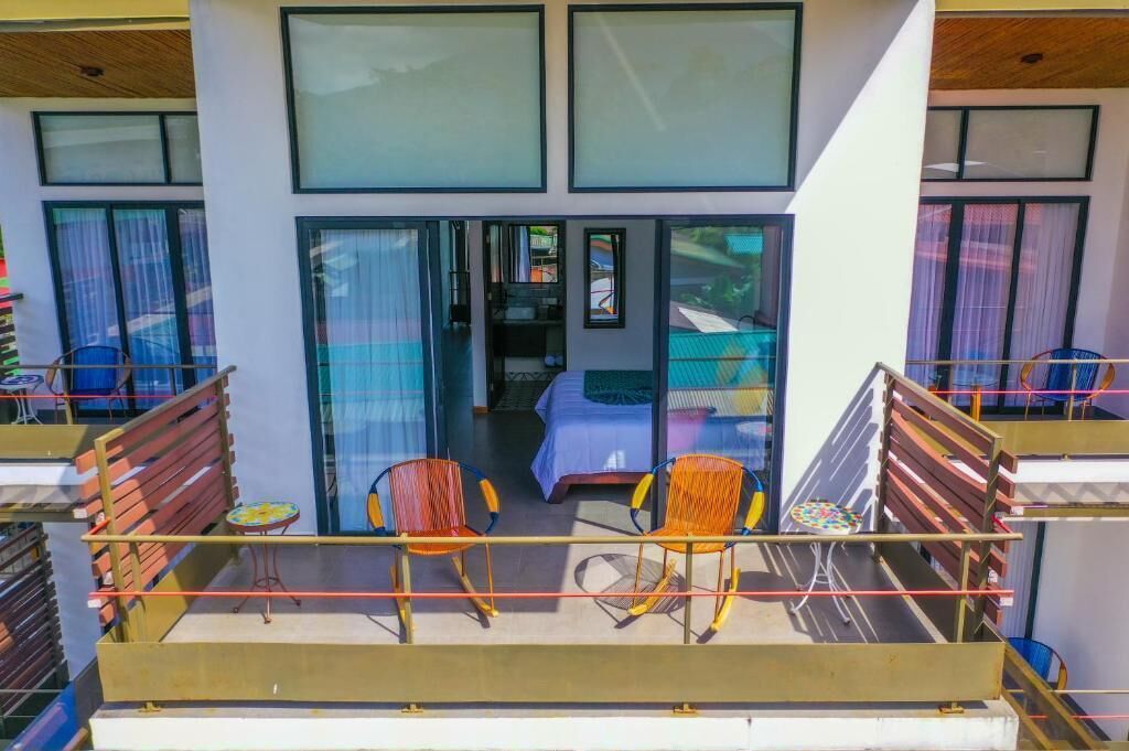 A balcony with two chairs and a small table, overlooking a bedroom with a bed and a window.
