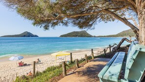 Une plage avec un banc en bois, un parasol à rayures et une personne assise sur une chaise longue.