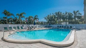 Ein rechteckiger Pool mit gewölbter Kante, umgeben von Liegestühlen und Sonnenschirmen, im Hintergrund Palmen und ein klarer, blauer Himmel.