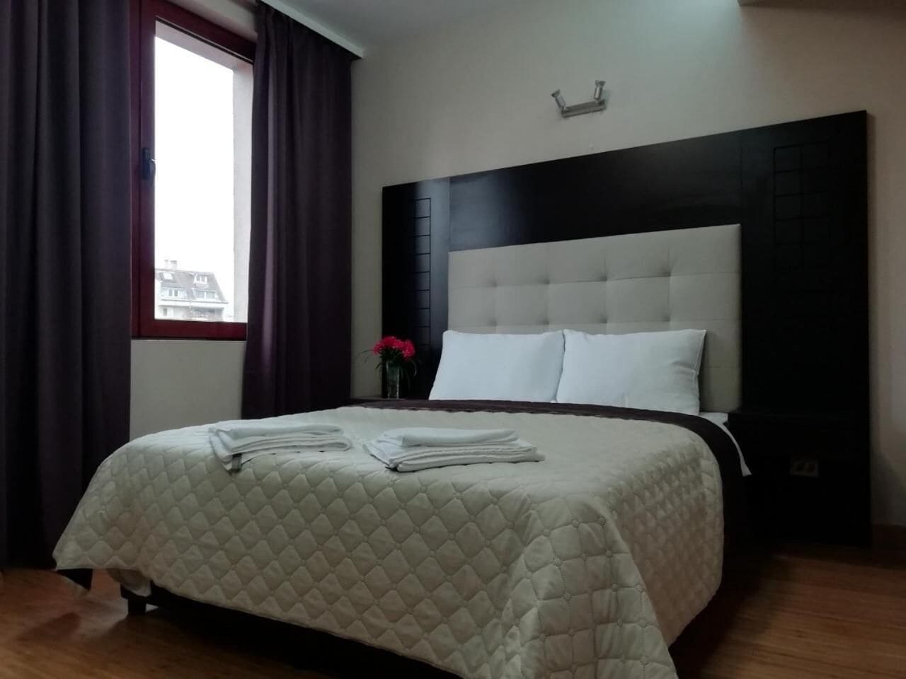 Una cama bien hecha con colcha acolchada, cabecera con panel oscuro y ventana con cortinas.
