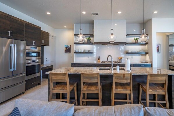 A modern kitchen with stainless steel appliances, wooden bar stools, and pendant lighting.