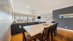 Un vaste espace de salle à manger avec une grande table et des chaises, une cuisine intégrée avec un mur de couleur gris foncé et un ventilateur de plafond.