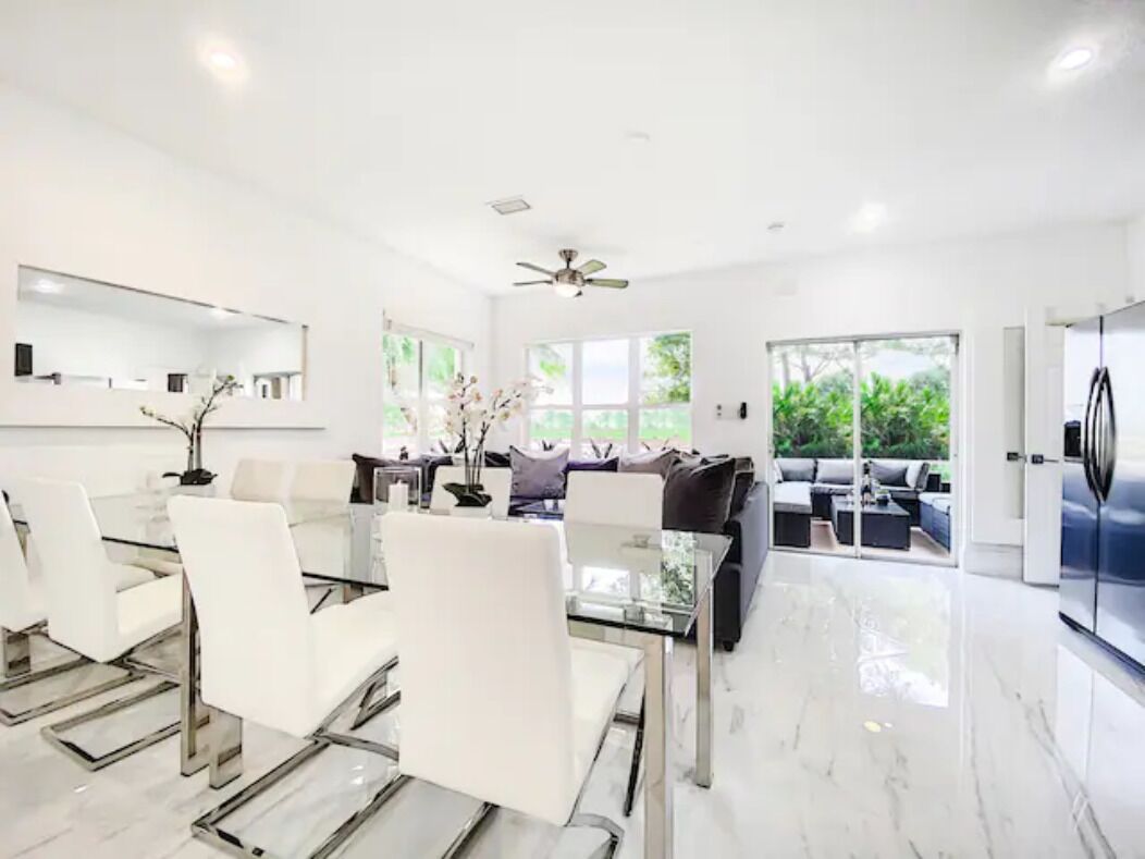 Un salon moderne avec une table à manger en verre, des chaises blanches, un canapé et un ventilateur de plafond.