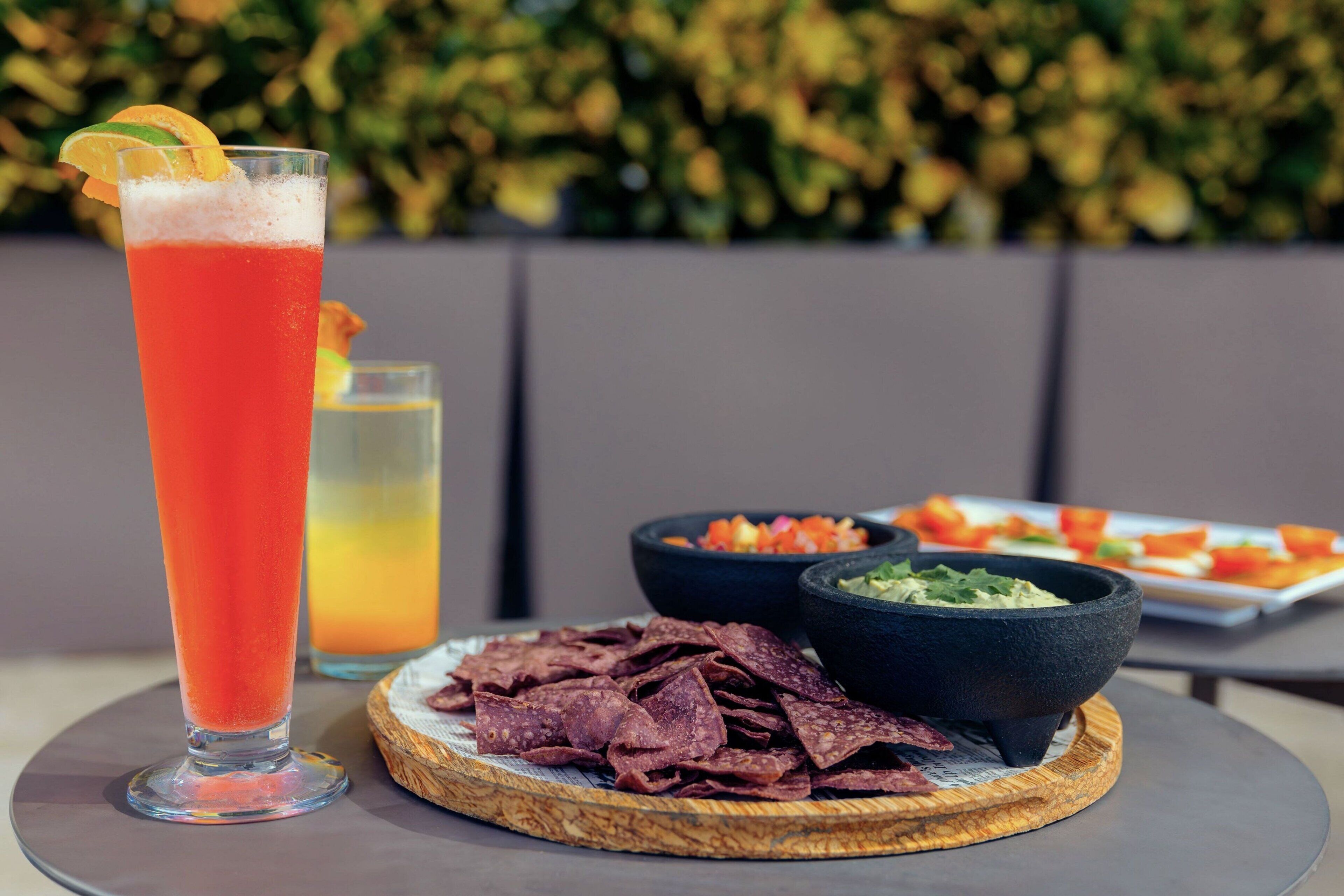 A table with a tall glass of red drink, a plate of chips, and two bowls of dips.