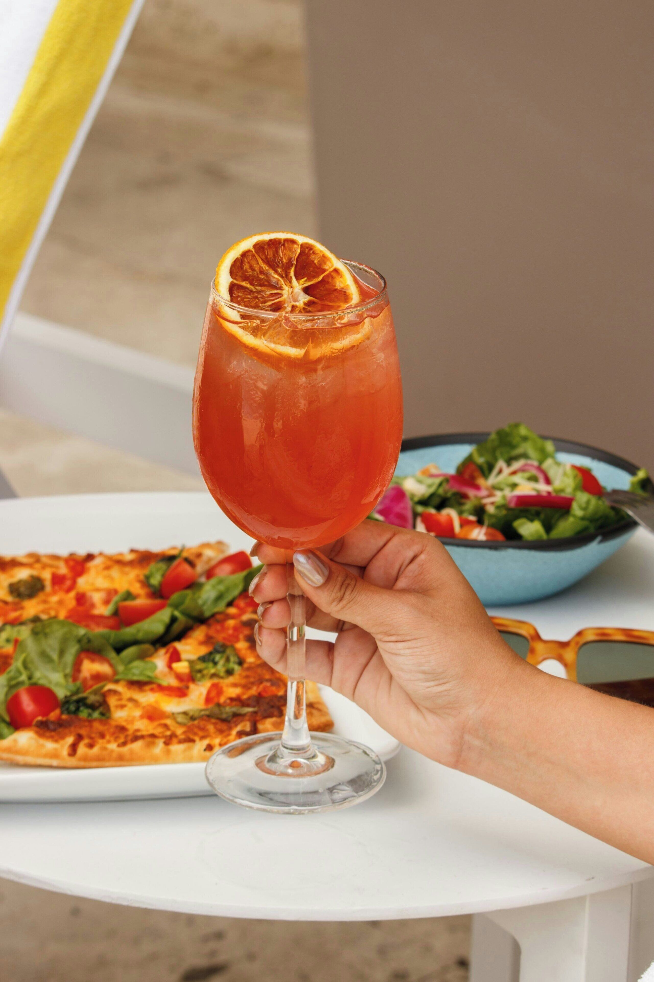 A hand holding a glass with a slice of orange on a white table. In the background, there is a pizza and a bowl of salad.