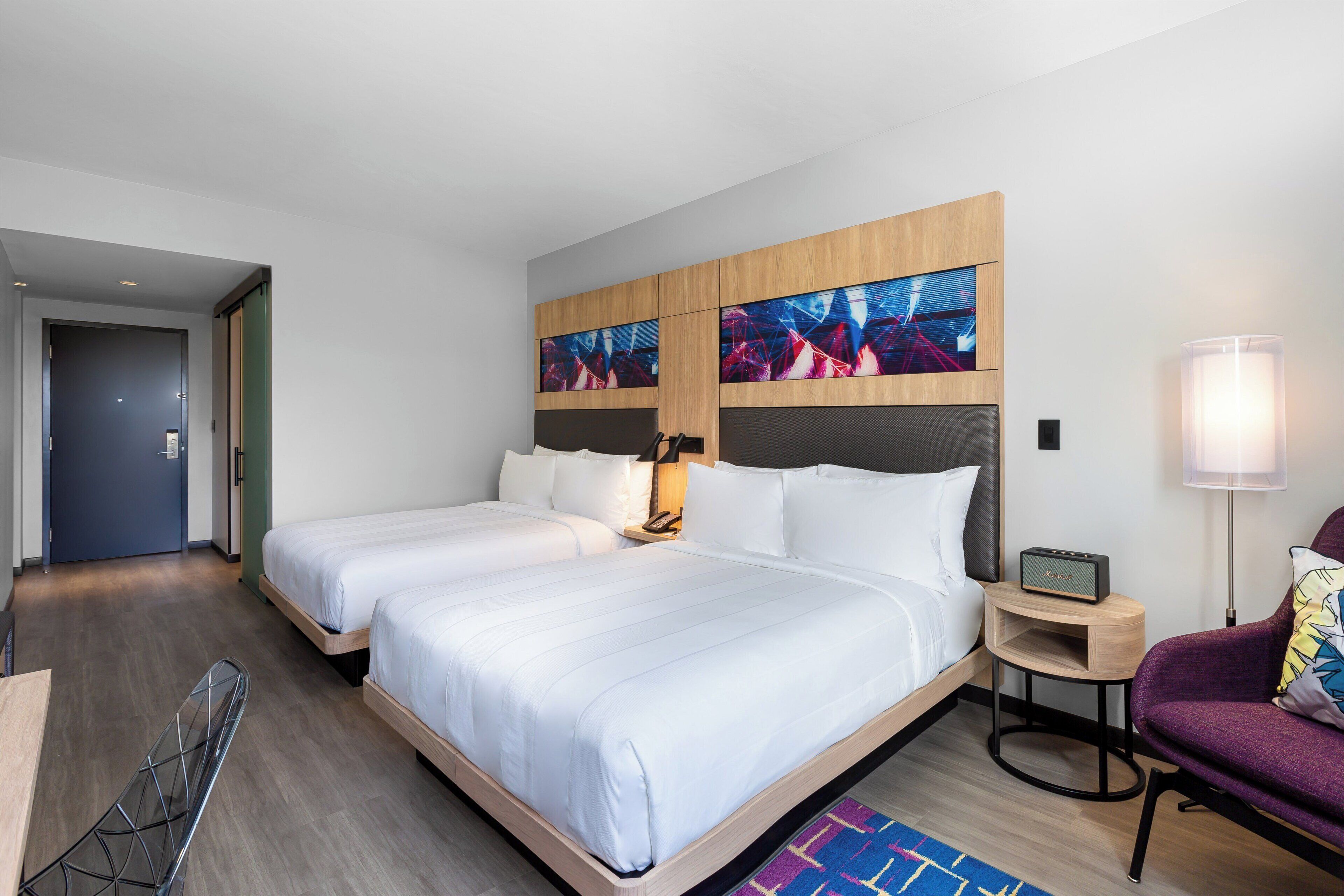 Habitación de hotel con dos camas, un escritorio, una silla, una lámpara y una obra de arte en la pared.