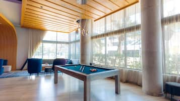 Una mesa de billar en una habitación luminosa con ventanas amplias, muebles modernos y techo de madera.