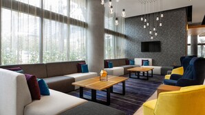 Un moderno espacio lounge con sofás, sillones y mesas, que cuenta con un amplio ventanal con cortinas, un televisor de pantalla plana montado en una pared texturizada e iluminación colgante.