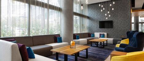 Un moderno espacio lounge con sofás, sillones y mesas, que cuenta con un amplio ventanal con cortinas, un televisor de pantalla plana montado en una pared texturizada e iluminación colgante.