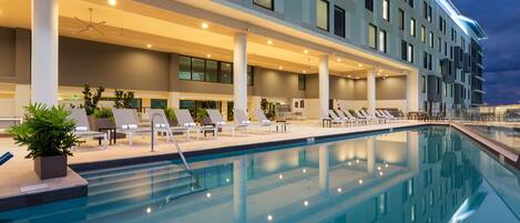 A modern hotel with a swimming pool, lounge chairs, and a covered area with plants.