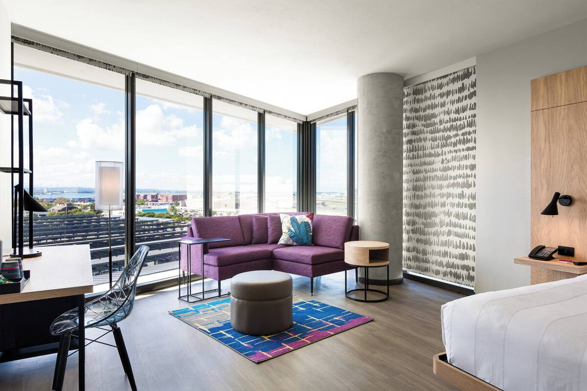 Una habitación de hotel moderna con un ventanal grande, un sofá morado, un escritorio de madera, una cama y una pared con estampado.
