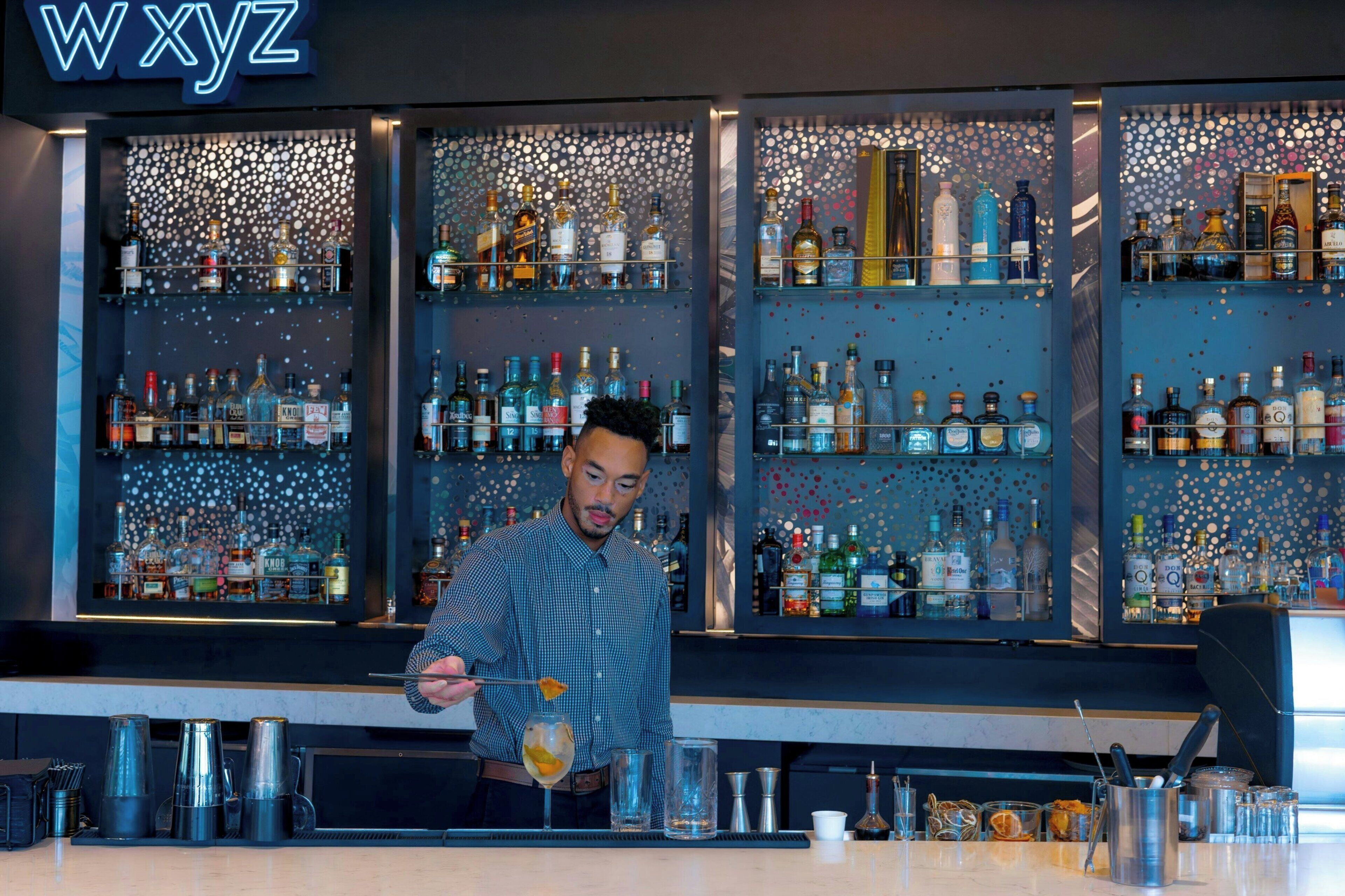 Ein Barkeeper hinter einer Bar mit Regalen voller Spirituosenflaschen und einem neonbeleuchteten Schild mit der Aufschrift 'wxyz'.