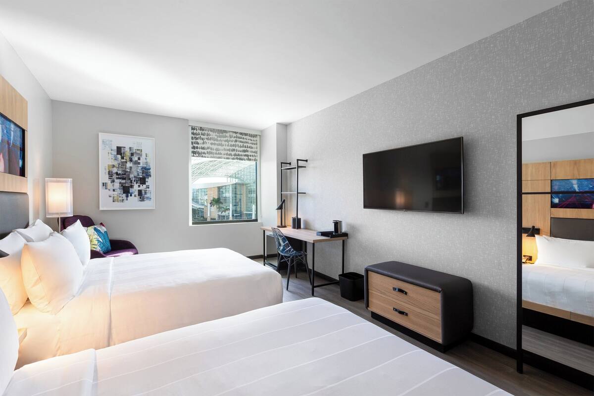Habitación de hotel con dos camas, un escritorio, una silla, un televisor y un ventanal grande.