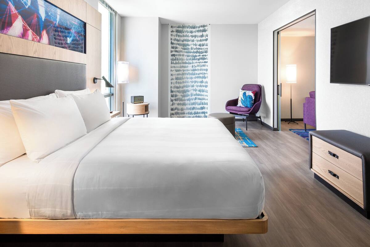 Una habitación de hotel moderna con una cama grande, un escritorio, una silla, un televisor y un ventanal amplio.
