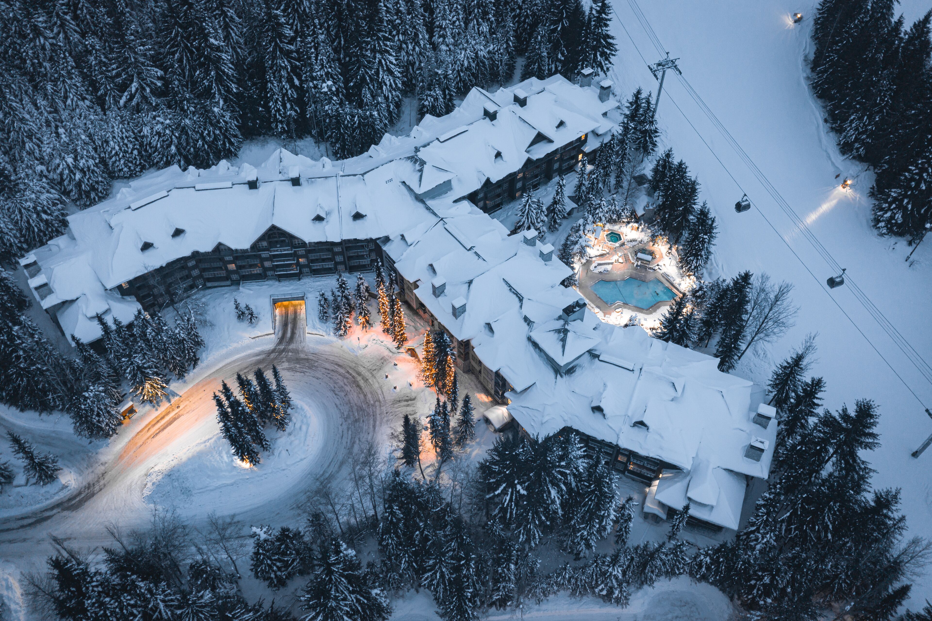 Eine Vogelperspektive zeigt einen schneebedeckten Ferienort mit einer Seilbahn, einem Poolbereich und einer kurvenreichen Straße.