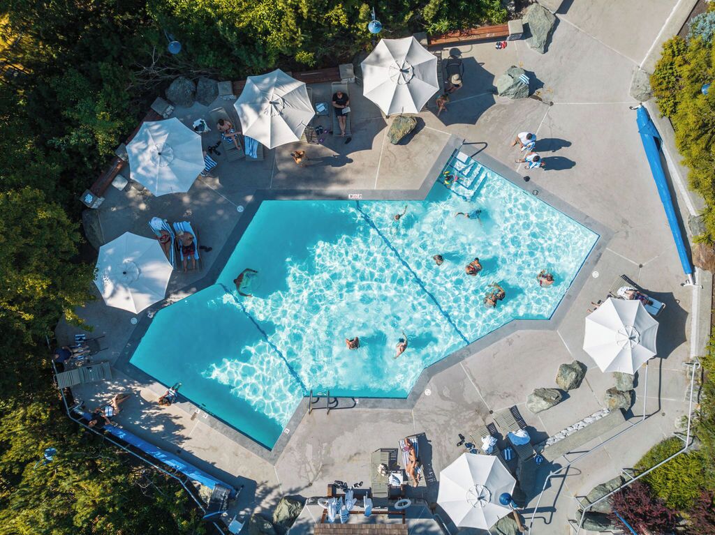 An aerial view of a swimming pool area with multiple sun loungers, umbrellas, and a few people swimming.