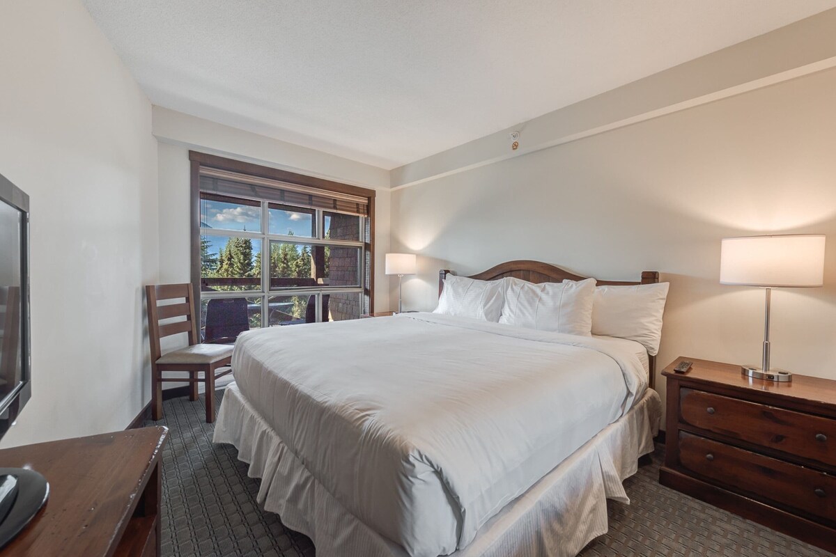 Ein Hotelzimmer mit einem großen Bett, einem Schreibtisch mit Computer, einem Sessel, einem Fenster mit Blick und einem Nachttisch mit Lampe.