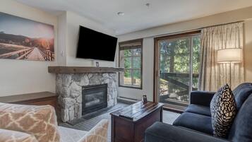A living room with a stone fireplace, a flat-screen TV mounted above it, and a large window offering a view of a wooden deck and trees.