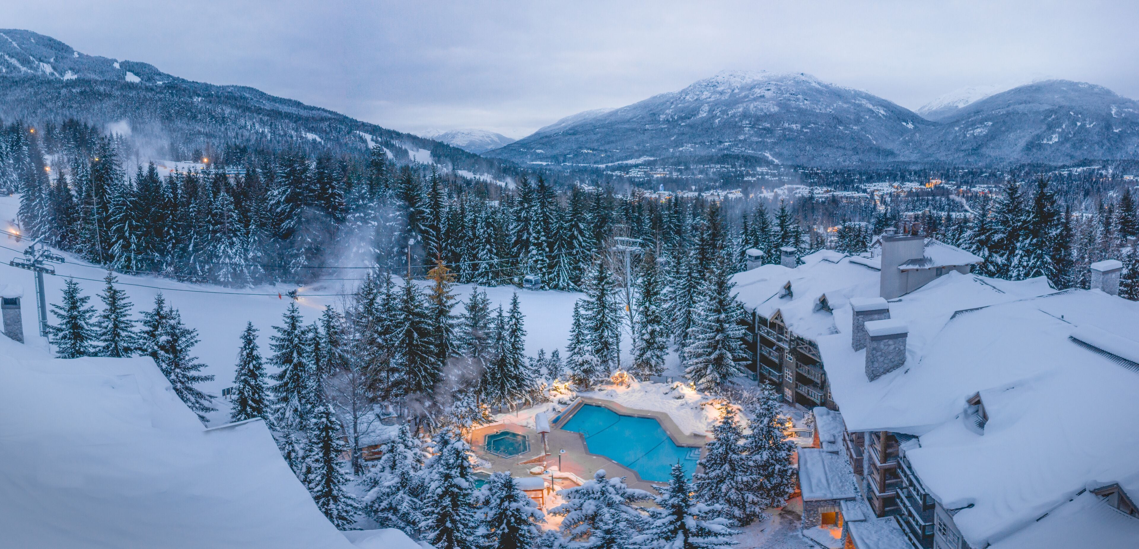 A resort with a heated outdoor pool surrounded by snow-covered trees and mountains in the background.