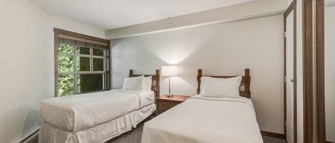 A hotel room with two single beds, a nightstand with a lamp, a window with a view of greenery, and a door.