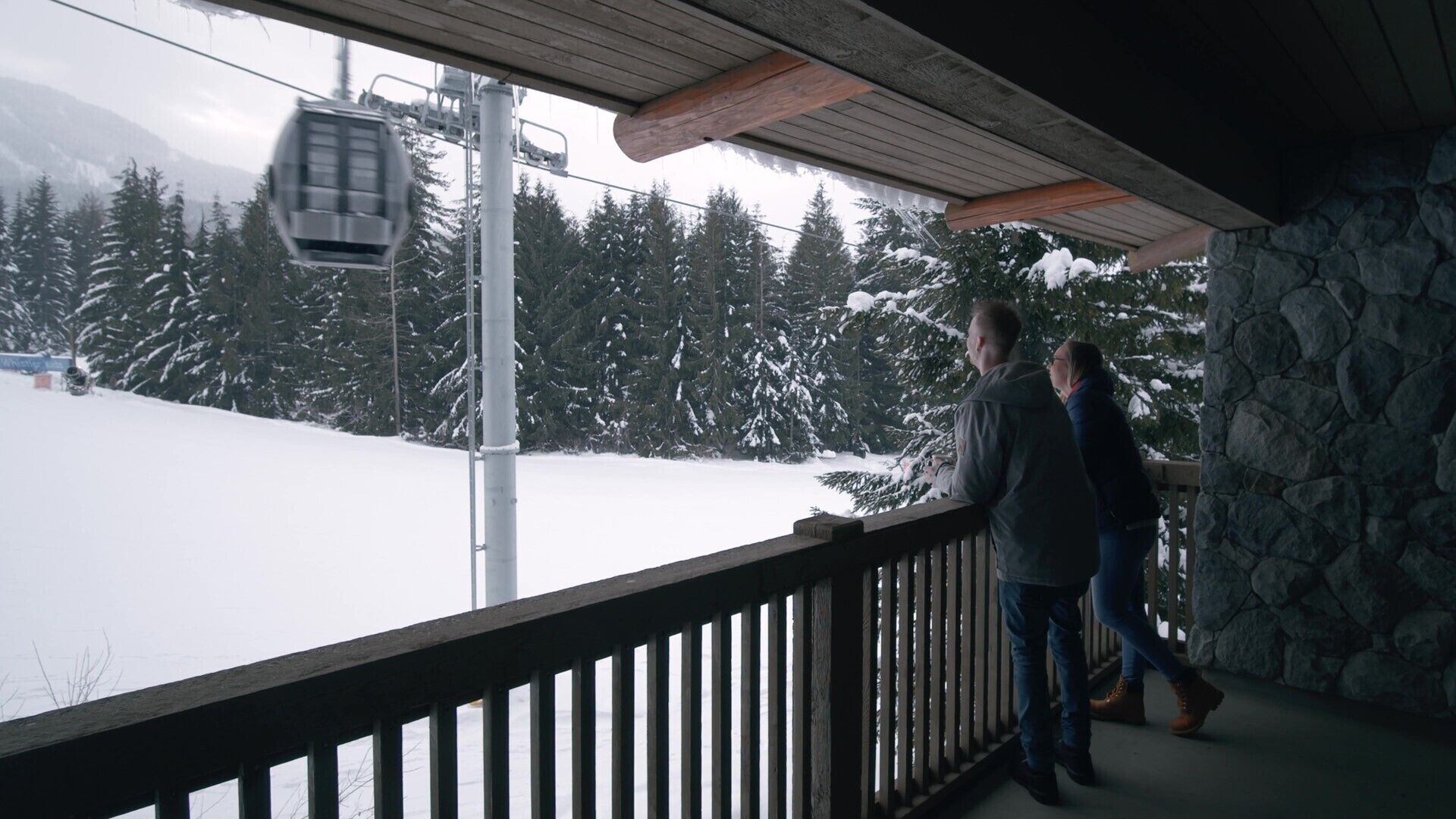 A wooden balcony with a stone wall, overlooking a snowy landscape with trees and a ski lift.