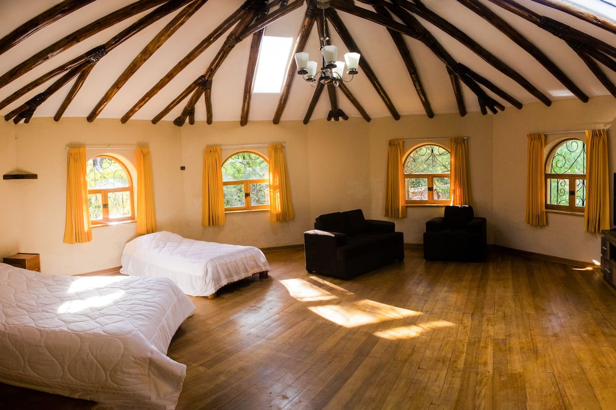Una habitación con vigas de madera, una cama, un sofá, un televisor y ventanas con cortinas.