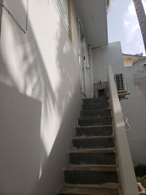 Un escalier menant à un bâtiment à la façade blanche, avec un petit appareil de climatisation et un pot de fleurs placé sur le palier.