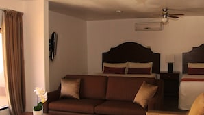 A hotel room with a brown sofa, a wooden coffee table, a small potted plant, a bed with a wooden headboard, and a ceiling fan. - Estancia Inn (Tecate)