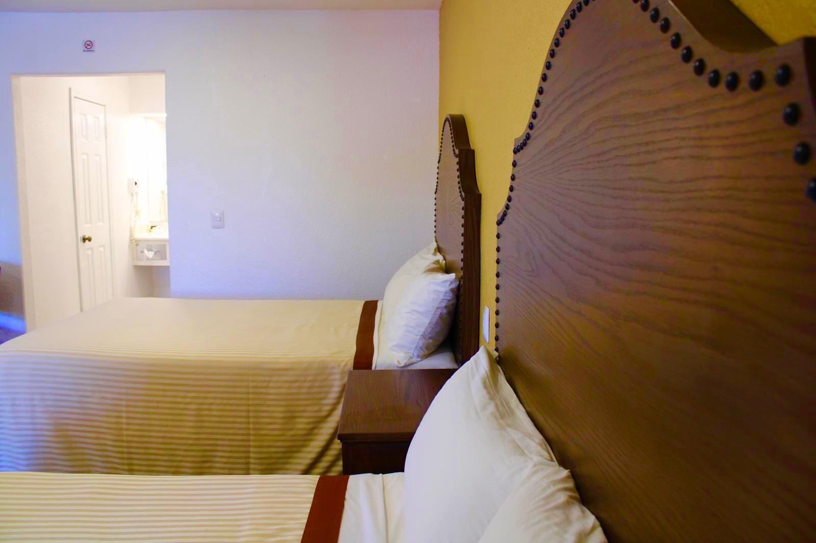 A hotel room with two beds, wooden headboards, and a small bedside table with a lamp.