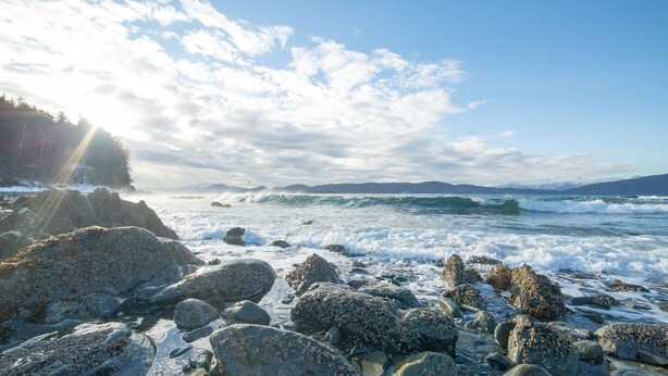Un rivage rocheux où les vagues s’écrasent contre les rochers, sous un ciel partiellement nuageux.