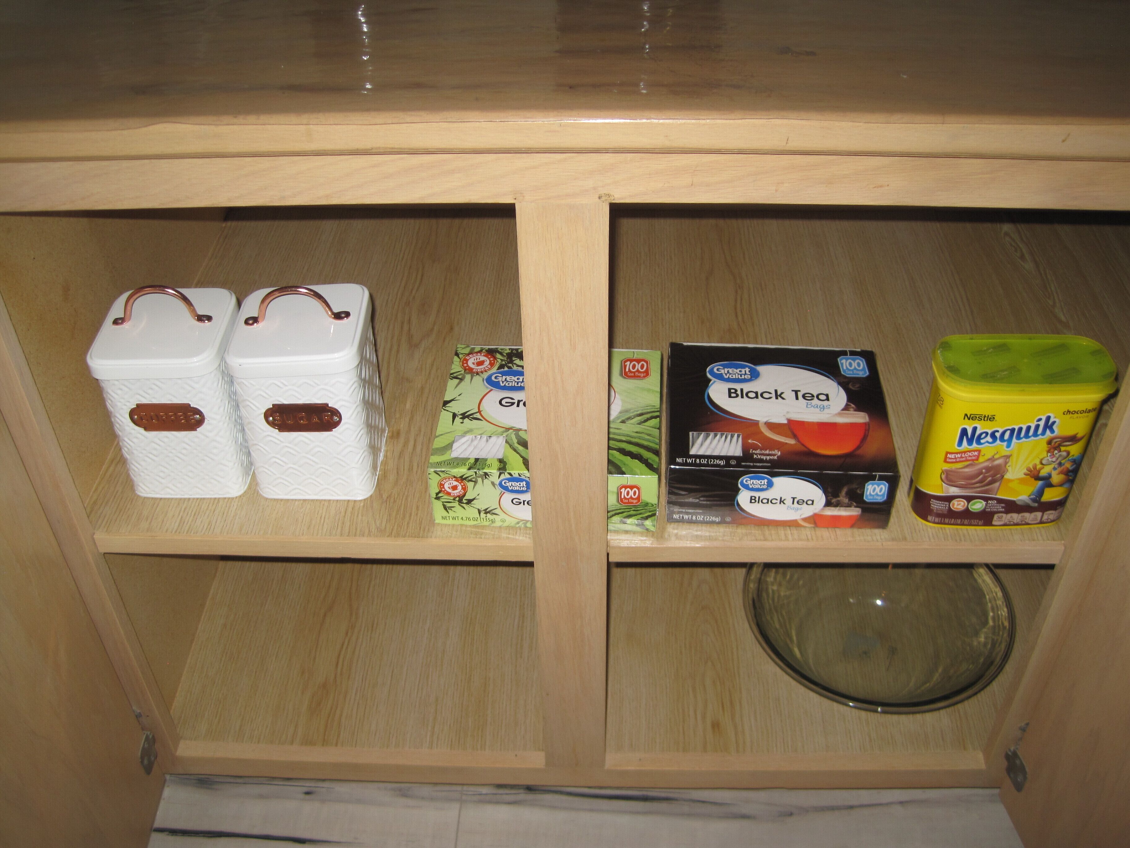 A wooden cabinet with two white containers labeled 'Coffee' and 'Sugar,' a box of 'Great Value Black Tea Bags,' and a 'Nestlé Nesquik' chocolate milk carton.